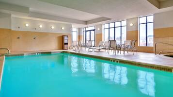 An indoor swimming pool with clear water, surrounded by lounge chairs and tables, and large windows allowing natural light.