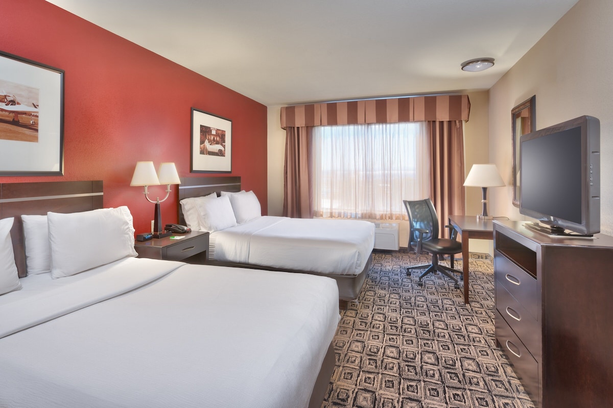 Quarto de hotel com duas camas, uma escrivaninha com cadeira, televisão e obras de arte nas paredes.