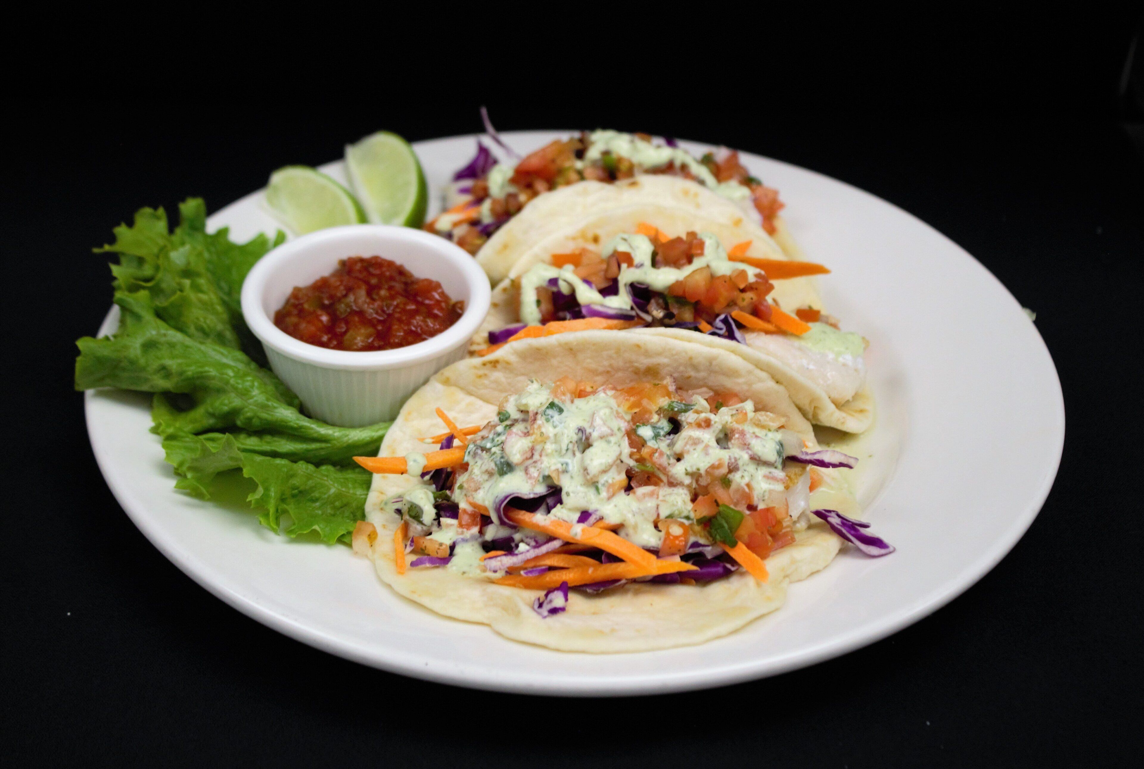 Three tacos on a plate with a side of lettuce, a small bowl of salsa, and a lime wedge.