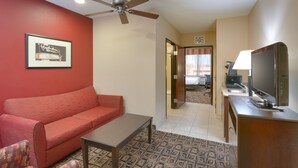 A hotel room with a red sofa, a small table, a ceiling fan, a television, and a kitchenette.