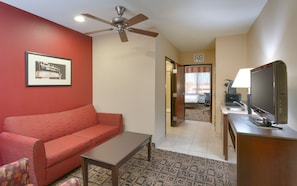 A hotel room with a red sofa, a small table, a ceiling fan, a television, and a kitchenette.