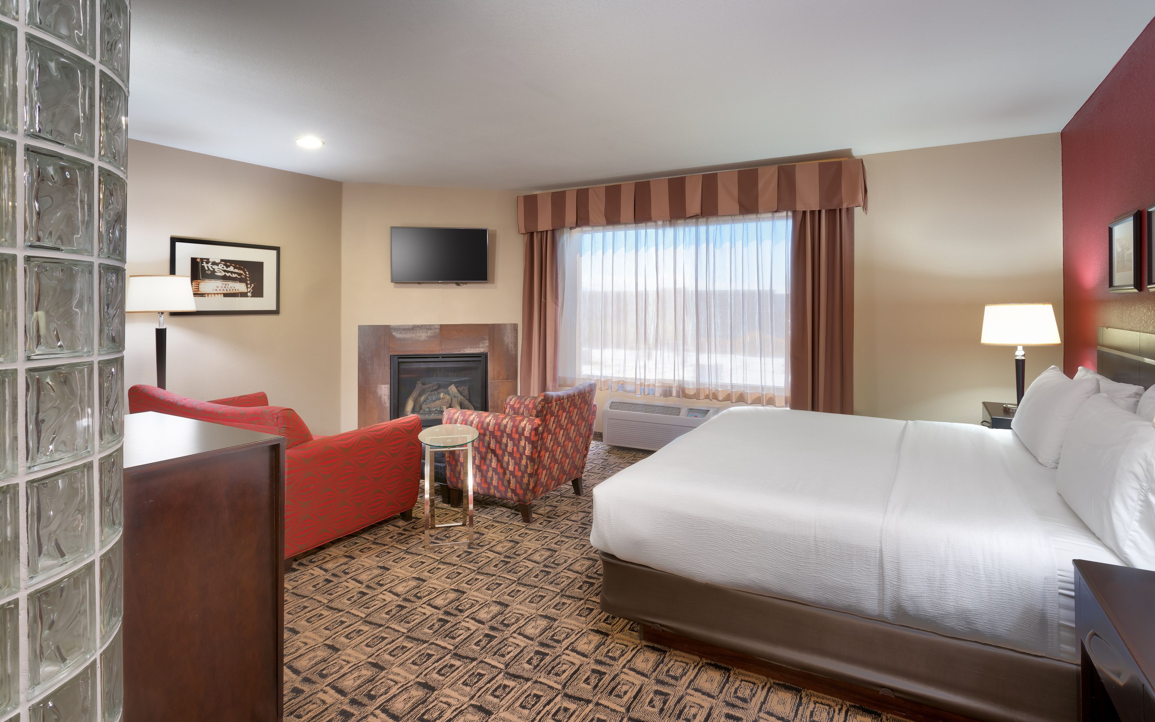 A hotel room with a large bed, a small sofa, a chair, a fireplace, and a television on the wall.