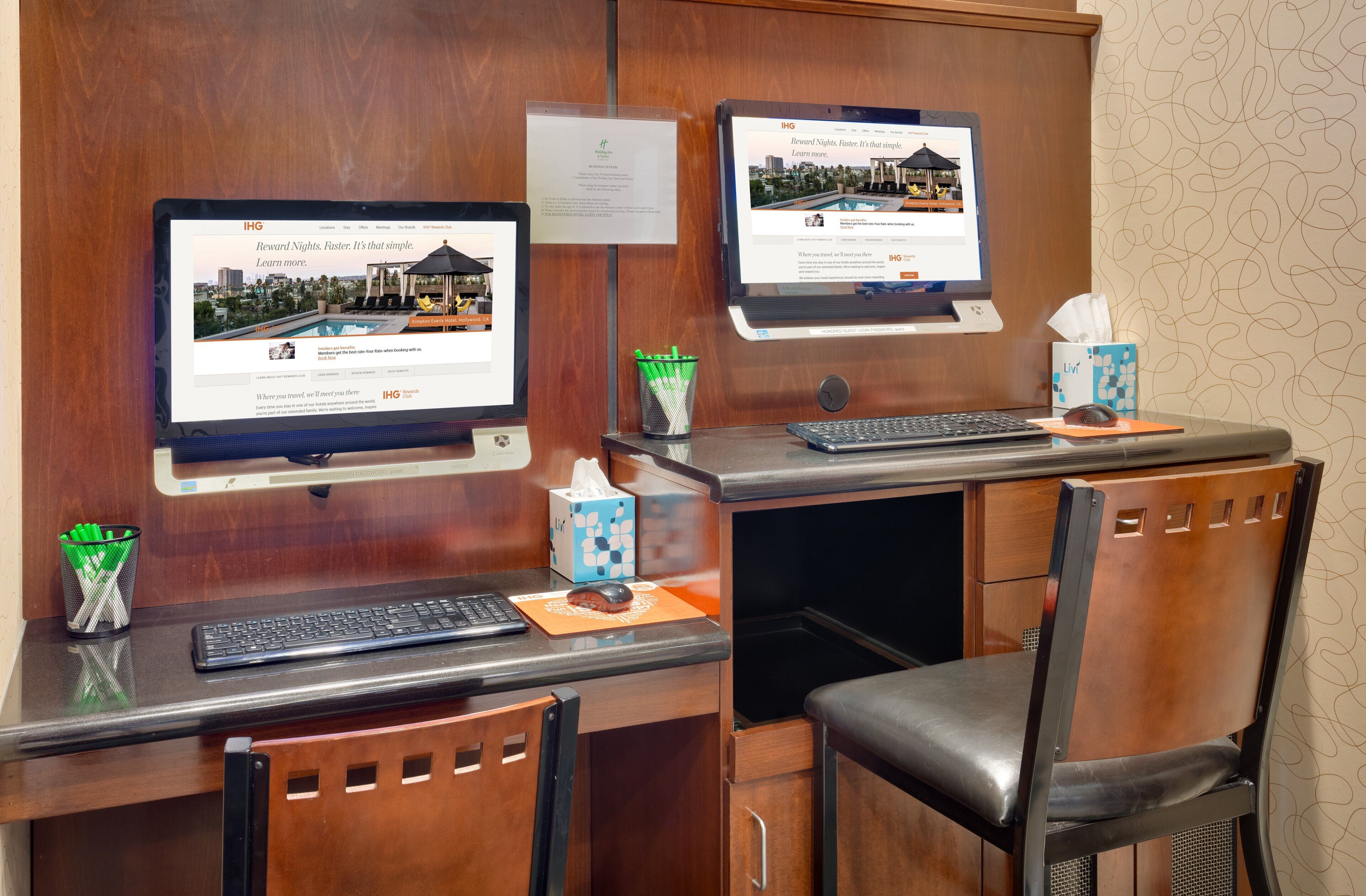 Un mostrador de recepción de hotel con dos monitores mostrando la página web del hotel, un teclado, un ratón, una caja de pañuelos y un portalápices.