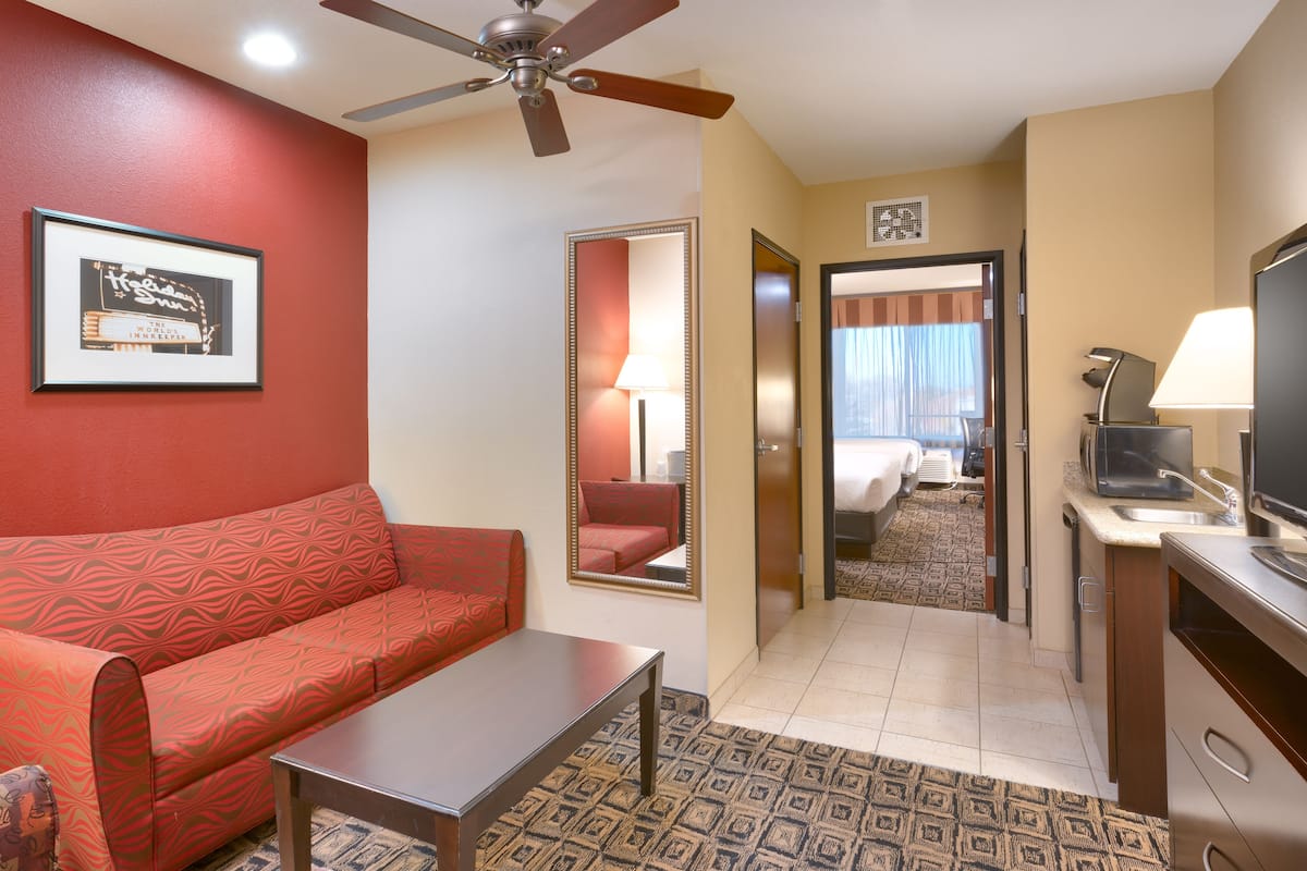 Une chambre d’hôtel avec un canapé rouge, une petite table, un ventilateur de plafond, un miroir, une télévision et une salle de bain visible à travers une porte ouverte.