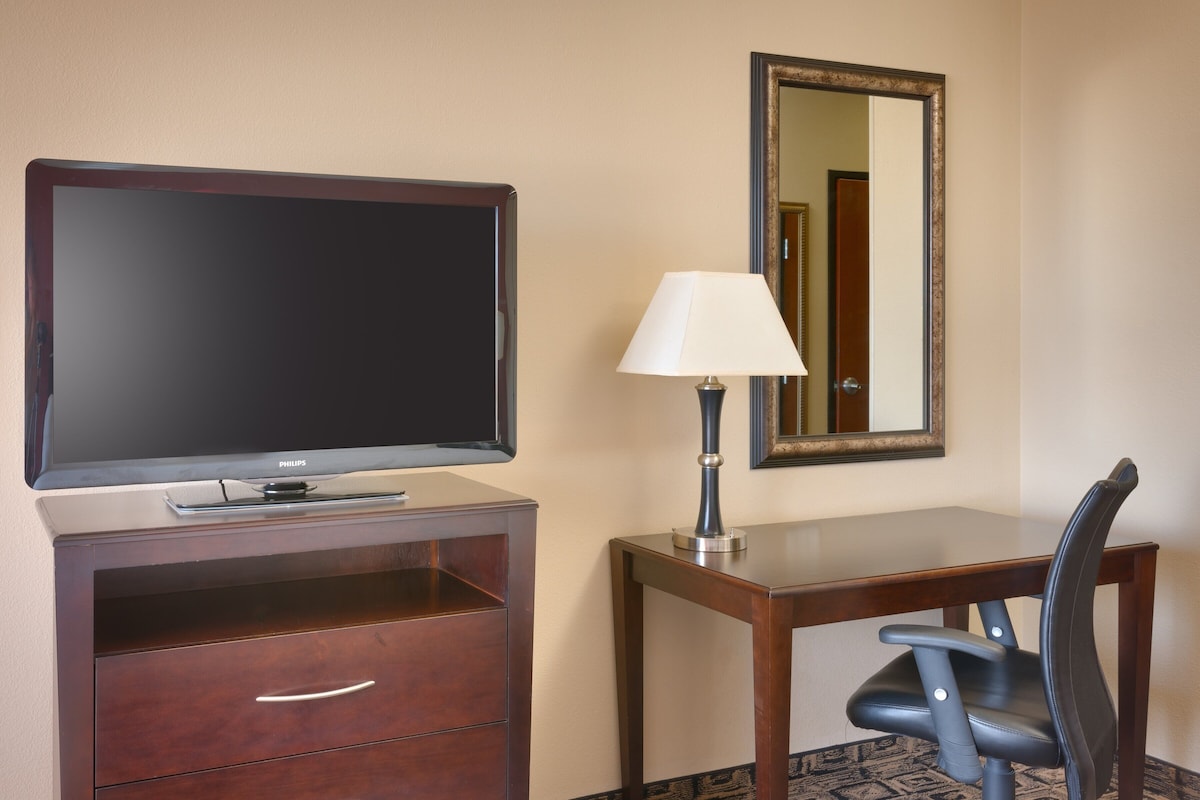 Quarto de hotel com televisão de tela plana, escrivaninha com luminária, cadeira e espelho na parede.