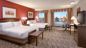 Quarto de hotel com duas camas, uma escrivaninha, uma cadeira, televisão e janelas amplas com cortinas.