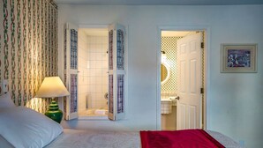 A bedroom with a bed, a nightstand, a green lamp, a red throw blanket, a bathroom with a bathtub, and a door leading to another room. - Auld Holland Inn (Oak Harbor)