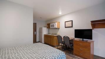 A compact hotel room with a bed, a kitchenette, a desk with a chair, and a television.