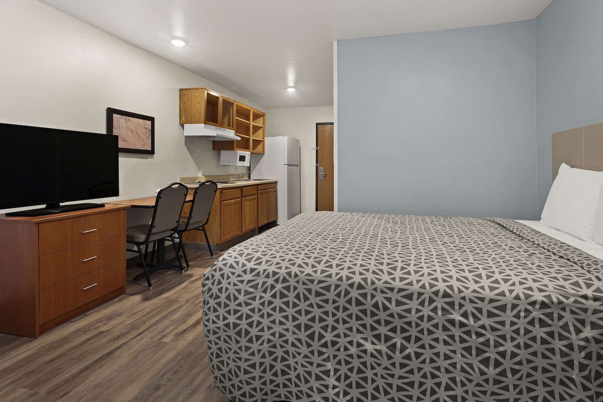 A bedroom with a bed, a desk with a computer, a chair, a TV, and a kitchenette.