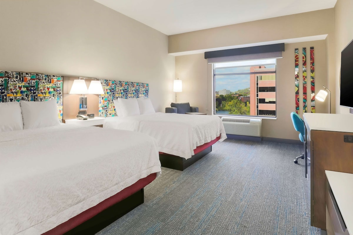 Quarto de hotel com duas camas, uma escrivaninha, uma cadeira, uma televisão e uma janela com vista para a cidade.