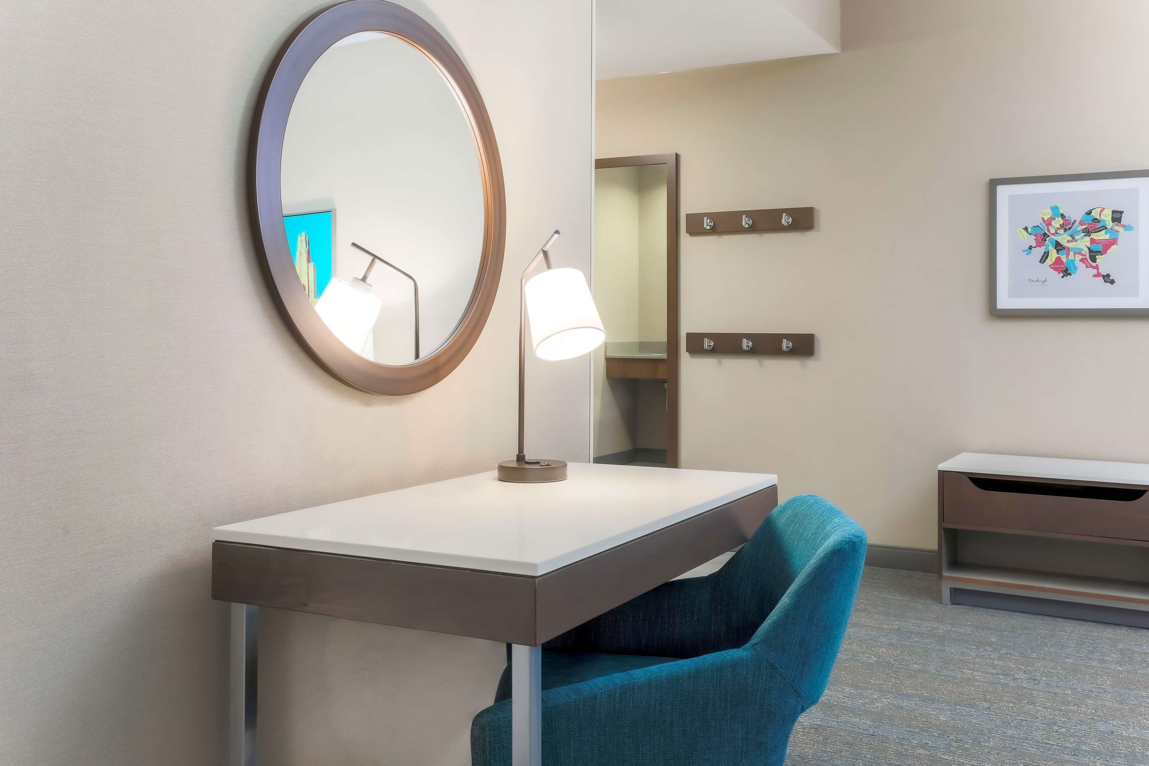 Ett modernt hotellrum med en rund spegel, ett skrivbord med en lampa, en blå stol och ett inramat konstverk på väggen.