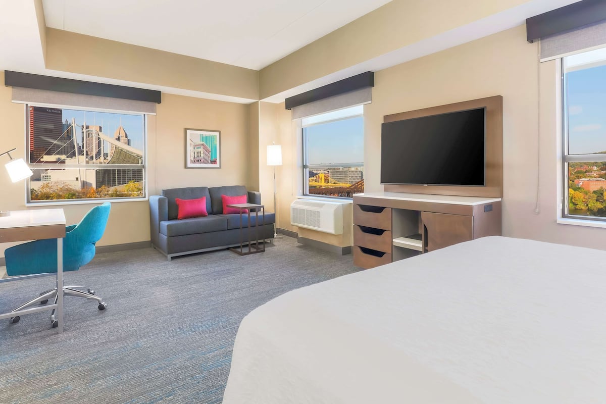 Quarto de hotel com cama, escrivaninha, cadeira, sofá com almofadas, televisão e amplas janelas com vista para a cidade.