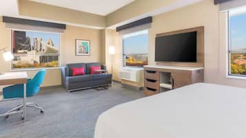 Ein Hotelzimmer mit Bett, Schreibtisch, Stuhl, einer Couch mit Kissen, einem Fernseher und großen Fenstern mit Blick auf die Stadt.