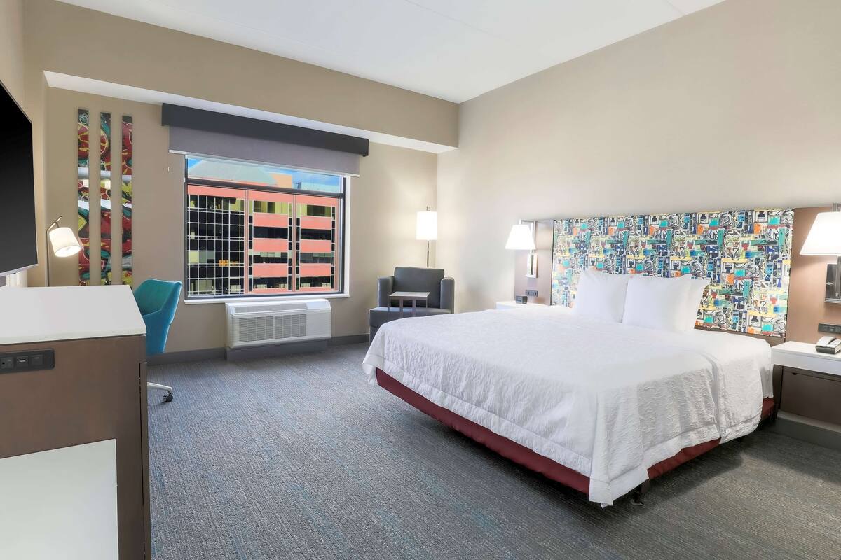 Quarto de hotel com uma cama grande, uma escrivaninha, uma cadeira, uma televisão e uma janela com vista para edifícios.