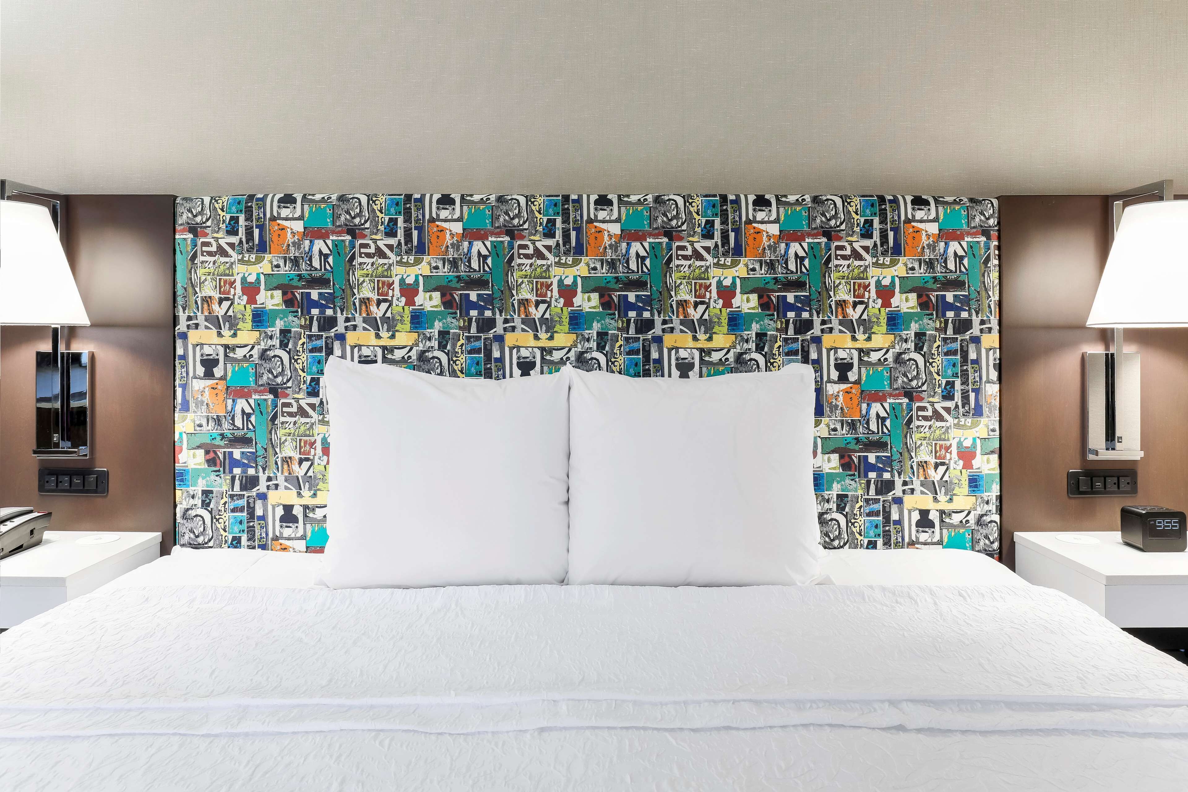 A hotel room with a large bed, white bedding, a colorful abstract headboard, and two bedside lamps.