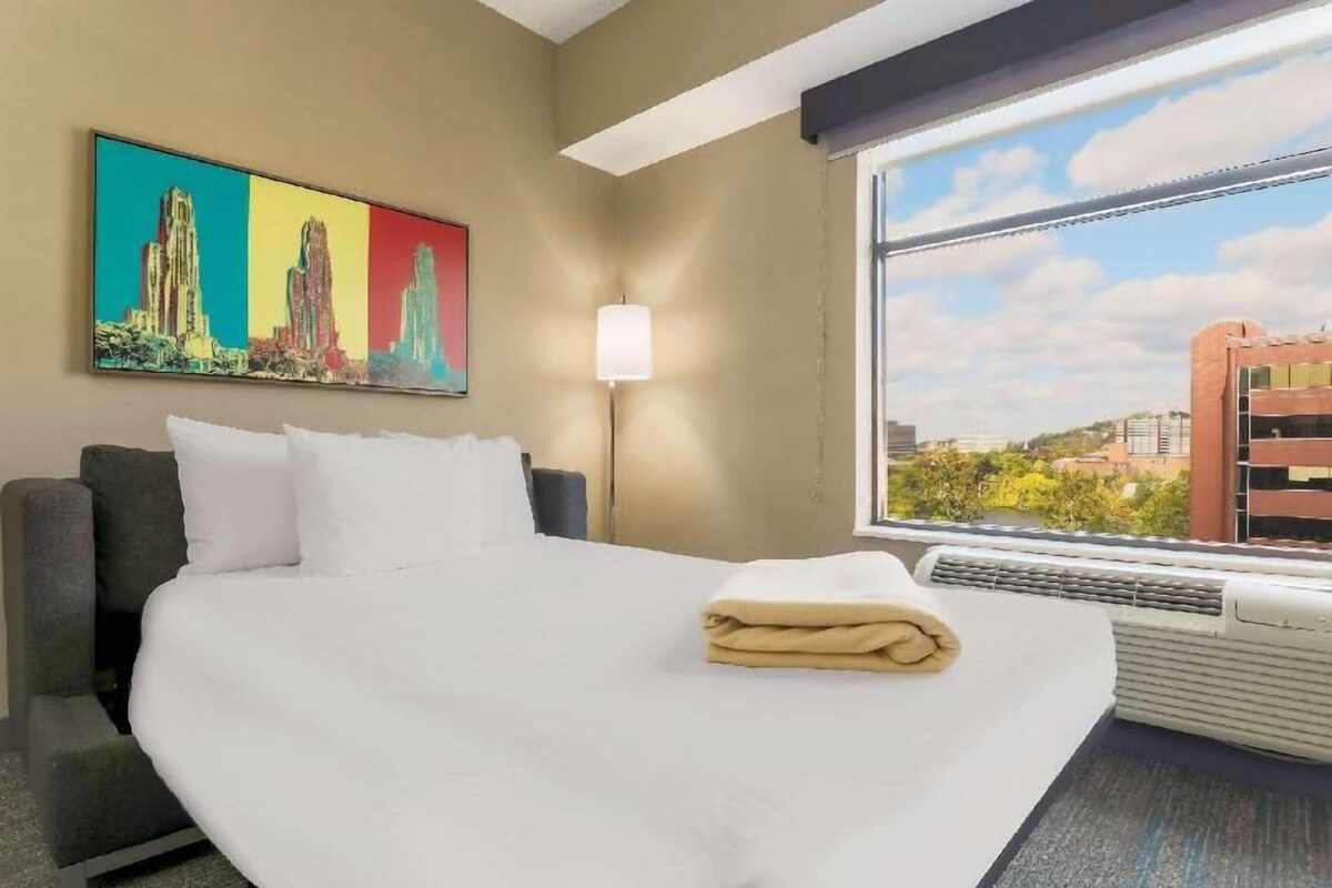 A hotel room with a bed, a large window with a city view, a painting on the wall, and a lamp.