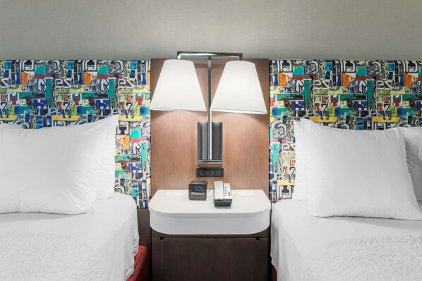A hotel room with two beds, a bedside table with a lamp, a clock, and a phone. The walls are decorated with colorful abstract patterns.