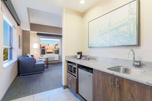 Cocina compacta con microondas integrado, lavavajillas y un lavabo. En la zona de estar contigua hay un sofá gris y una obra de arte grande con marco.