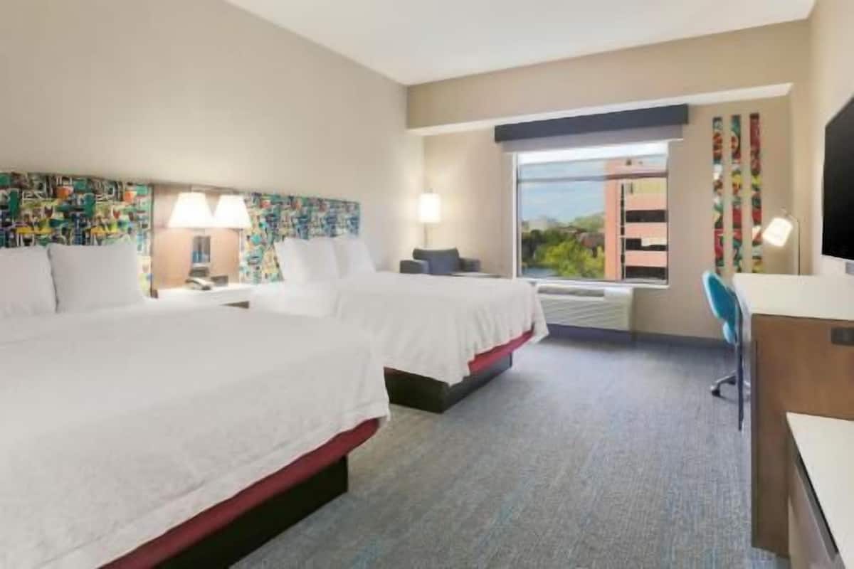Quarto de hotel com duas camas, uma escrivaninha, uma cadeira, uma janela com vista e uma cabeceira colorida.