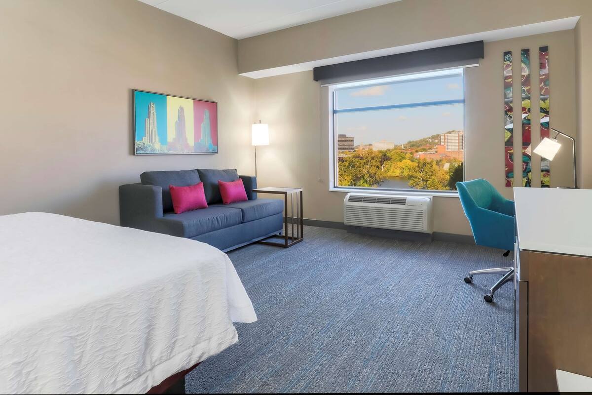 Quarto de hotel com cama, sofá com almofadas, escrivaninha, cadeira, janela grande com vista para a cidade e um quadro na parede.
