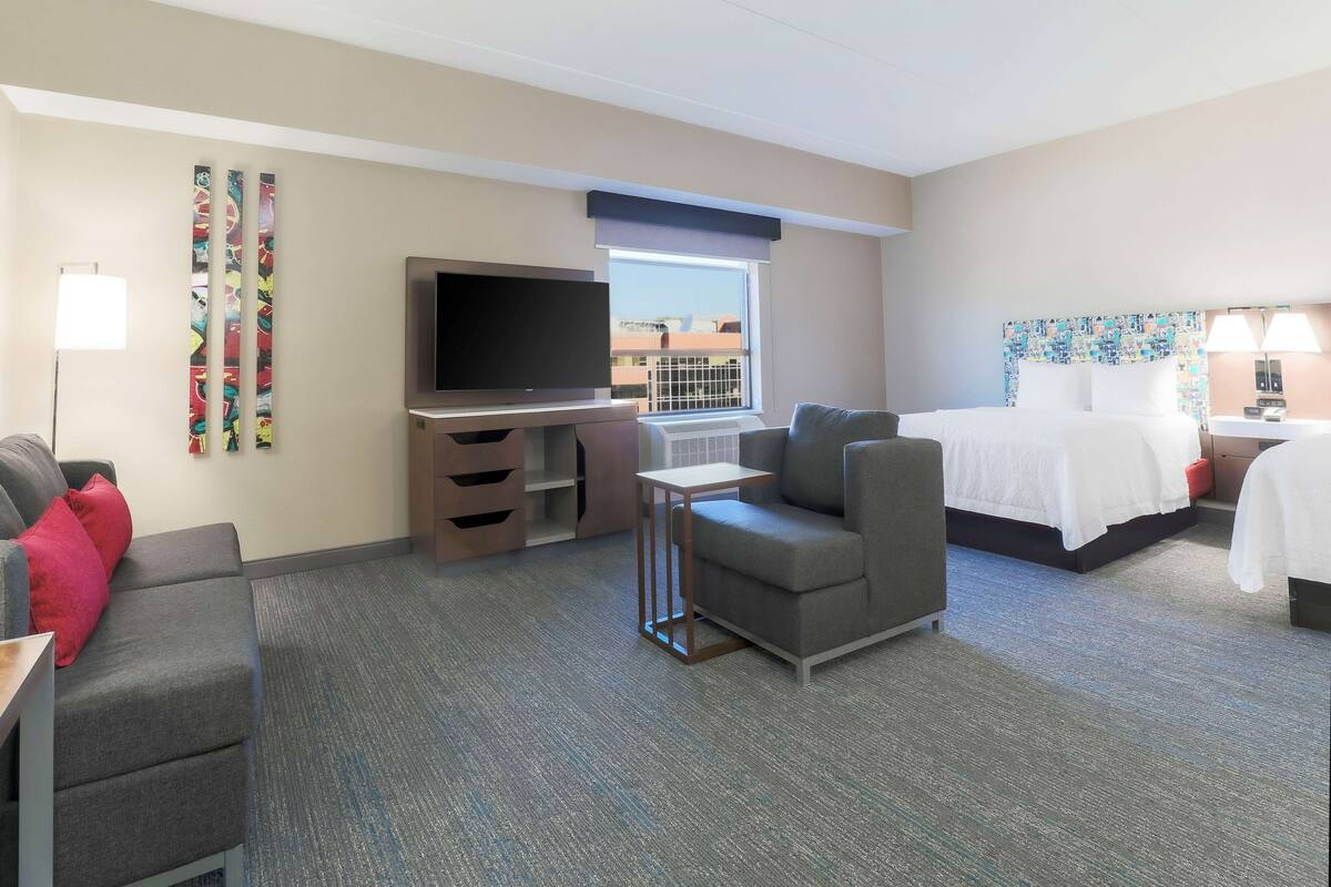 Quarto de hotel com cama, televisão, sofá, mesinha e janela com vista.