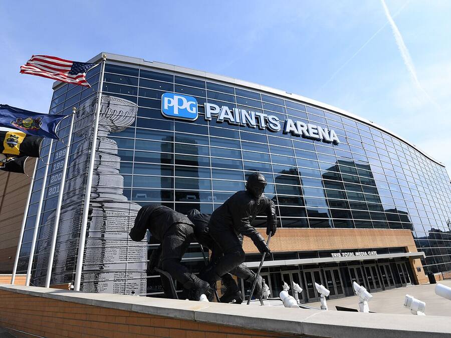 Exterior del PPG Paints Arena, con un amplio ventanal de vidrio, una estatua de un jugador de béisbol y una bandera estadounidense.
