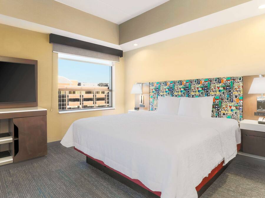 Quarto de hotel com uma cama grande, televisão de tela plana, escrivaninha e janela com vista para edifícios.