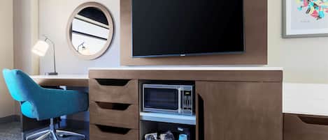 A hotel room with a flat-screen TV mounted on the wall, a blue office chair, a desk with drawers, and a framed artwork on the wall.