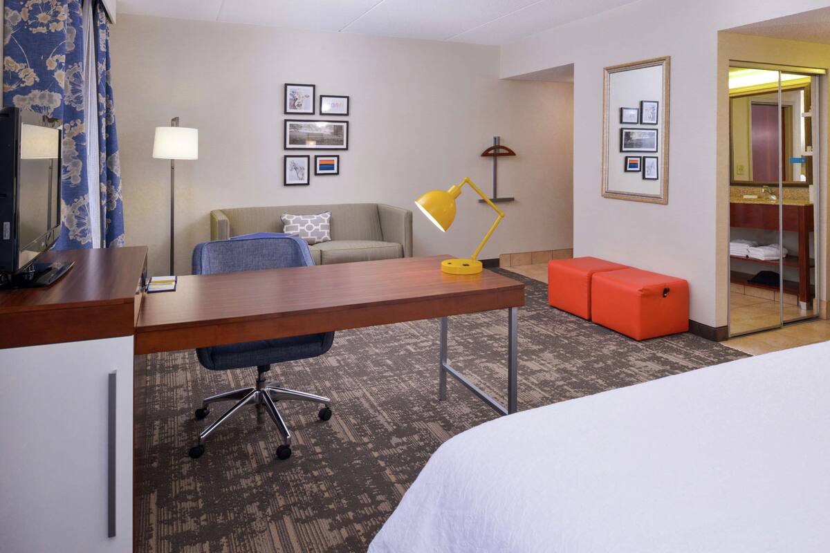 A hotel room with a wooden desk, a blue office chair, a bed, a red ottoman, a lamp, a sofa, and a mirror.