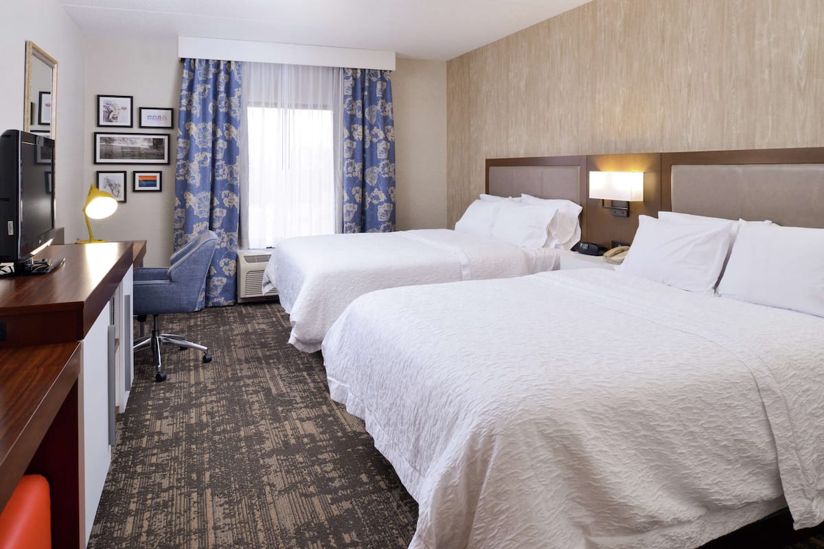 A hotel room with two beds, a desk, a chair, a desk lamp, and a window with curtains.