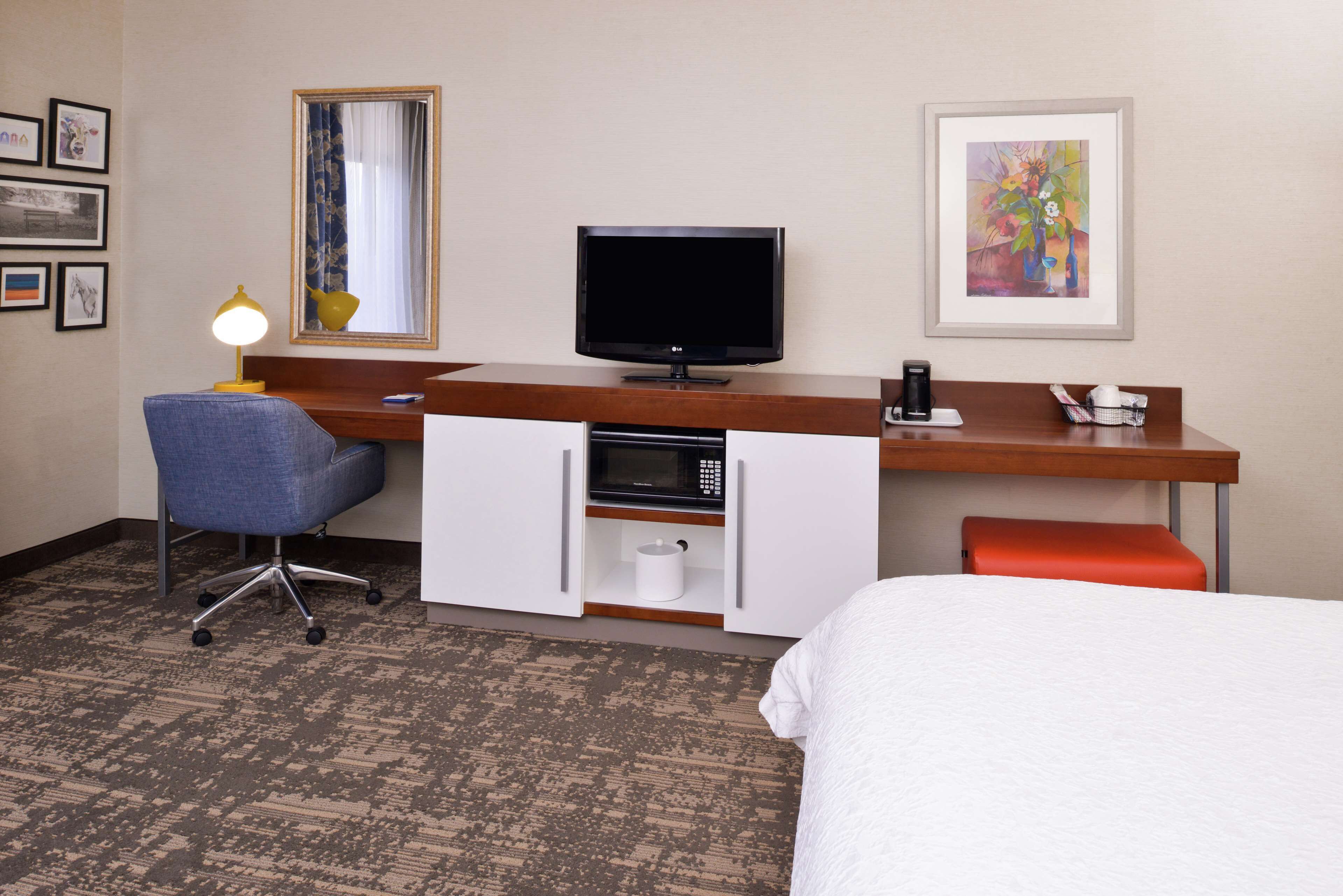 A hotel room with a bed, a desk with a computer and a TV, a chair, a microwave, and a coffee maker.