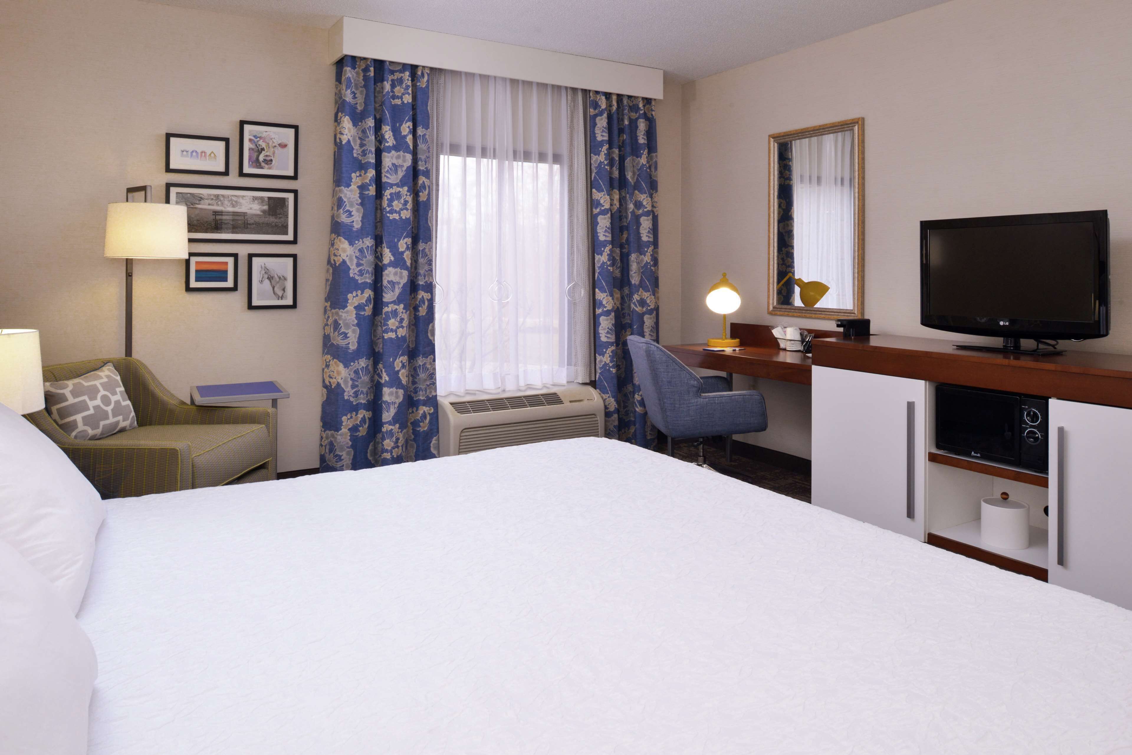 A hotel room with a bed, a desk with a television, a chair, a desk lamp, and a window with curtains.