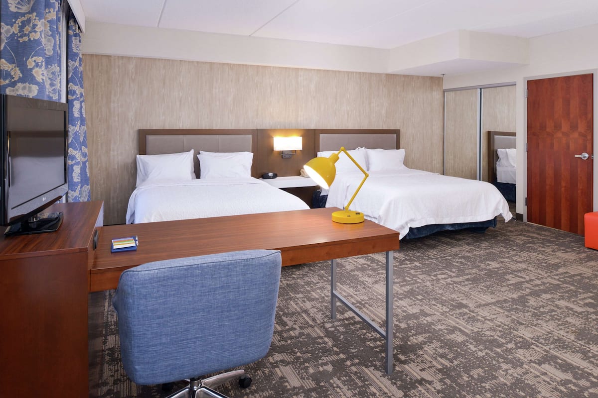 A hotel room with two beds, a desk with a chair, a TV, and a lamp.