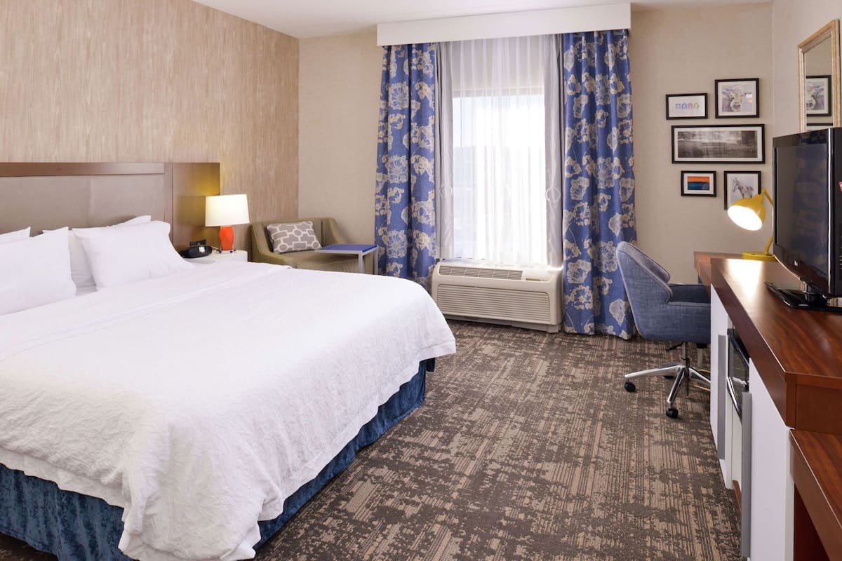 A hotel room with a large bed, a desk with a computer, a television, and a window with curtains.