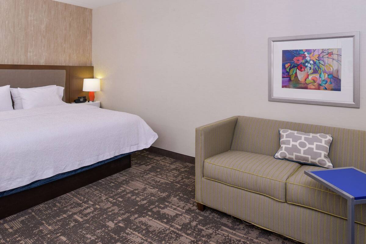 A hotel room with a bed, a nightstand with a lamp, a sofa, and a framed artwork on the wall.