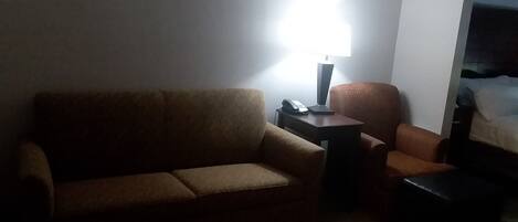 A dimly lit room with a brown sofa, a small table with a lamp, and a chair.