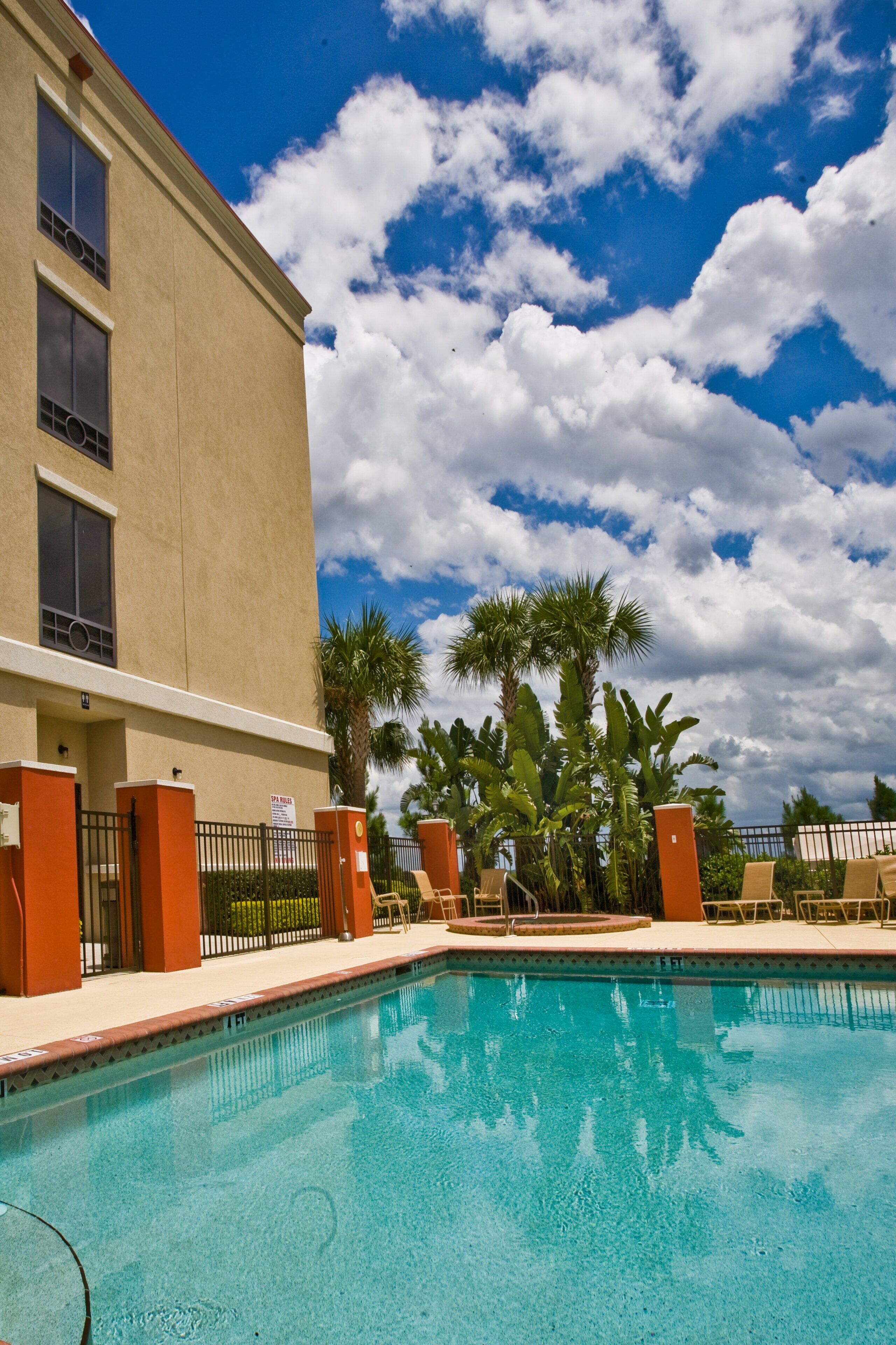 Una piscina con superficie de agua azul cristalina, rodeada de palmeras y un edificio de fachada color crema.