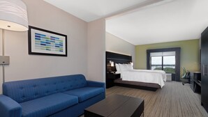 A hotel room with a blue sofa, a bed with white linens, a dark wooden coffee table, and a large window offering a view.