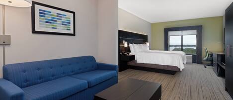 Ein Hotelzimmer mit blauem Sofa, einem Bett mit weißer Bettwäsche, einem dunklen Holztisch und einem großen Fenster mit Blick.