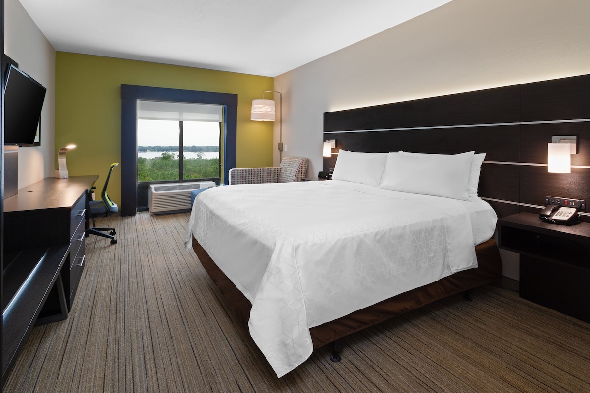 Habitación de hotel con una cama grande, un escritorio con computadora, una silla, un televisor y una ventana con vistas.