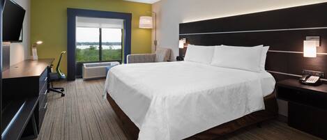 Quarto de hotel com uma cama grande, uma escrivaninha com computador, uma cadeira, uma televisão e uma janela com vista.