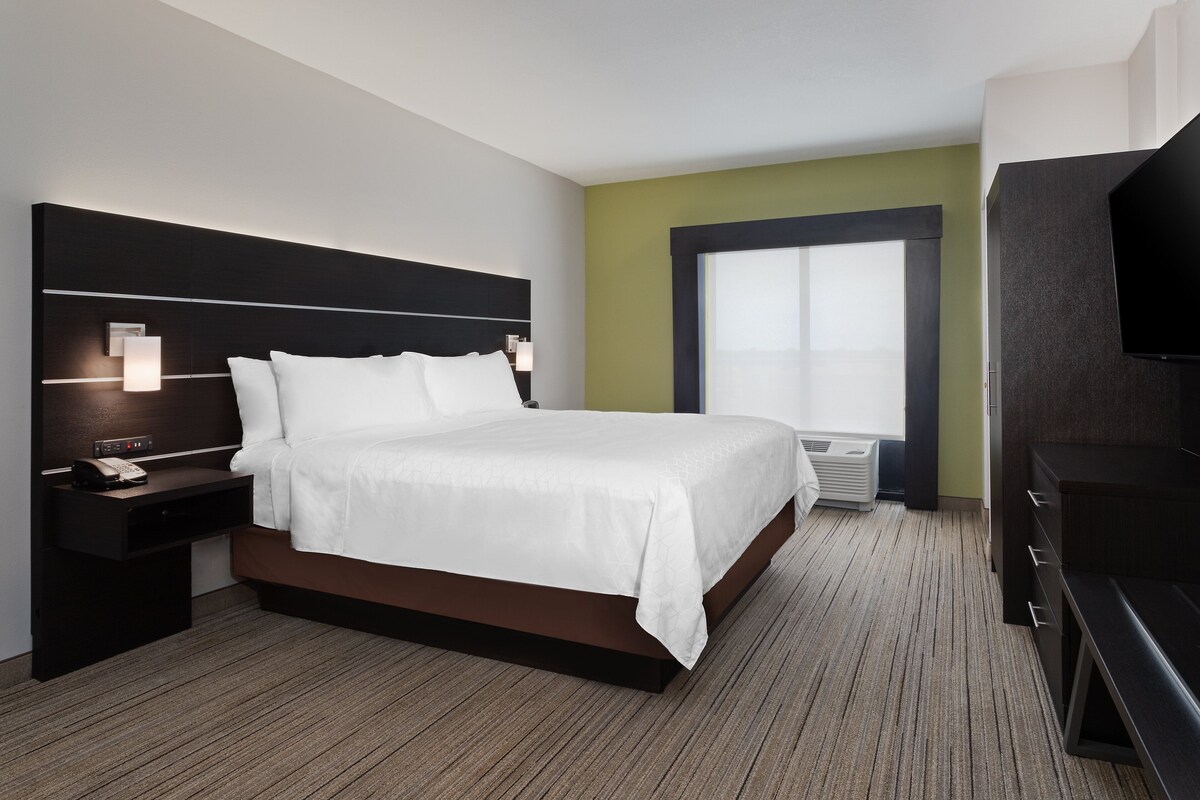 Una habitación de hotel moderna con una cama grande, mesitas de noche, un televisor de pantalla plana y un ventanal con persianas.