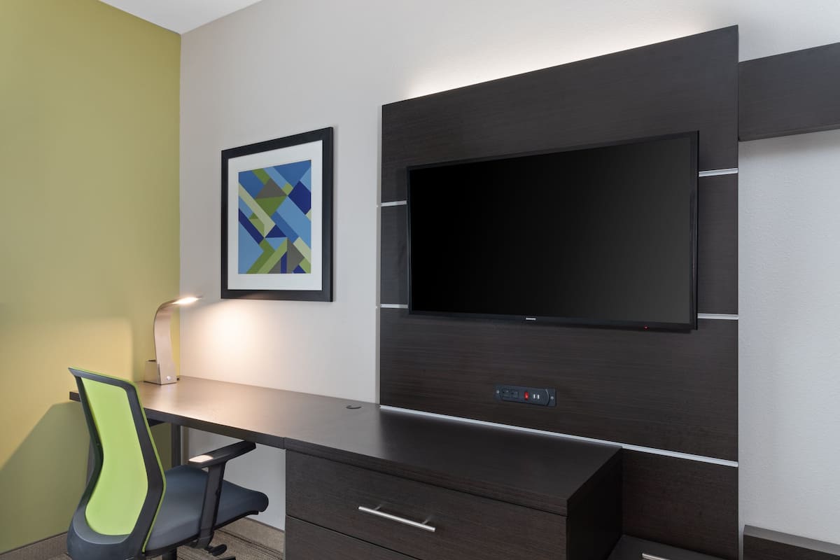 Habitación de hotel moderna con un escritorio de madera oscura, un televisor de pantalla plana montado en la pared, una silla verde y negra, y una obra de arte abstracta enmarcada en la pared.