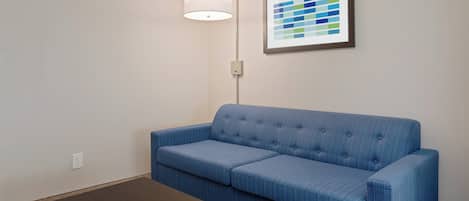Una stanza con un divano blu rivestito in tessuto, un tavolo di legno marrone, una lampada a parete e una tavolozza di colori incorniciata appesa al muro.