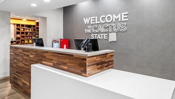 A reception desk with a wooden panel design, a wall-mounted sign reading "WELCOME TO THE CACTUS STATE," and a background view of a snack-filled display. - Hampton Inn & Suites Farmington (Farmington)