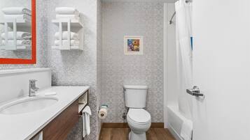 Ein Badezimmer mit einem weißen Waschbecken, einer Toilette und einer Dusche. Handtücher liegen auf einem Regal und ein Bild ist in einem Bilderrahmen an der Wand.