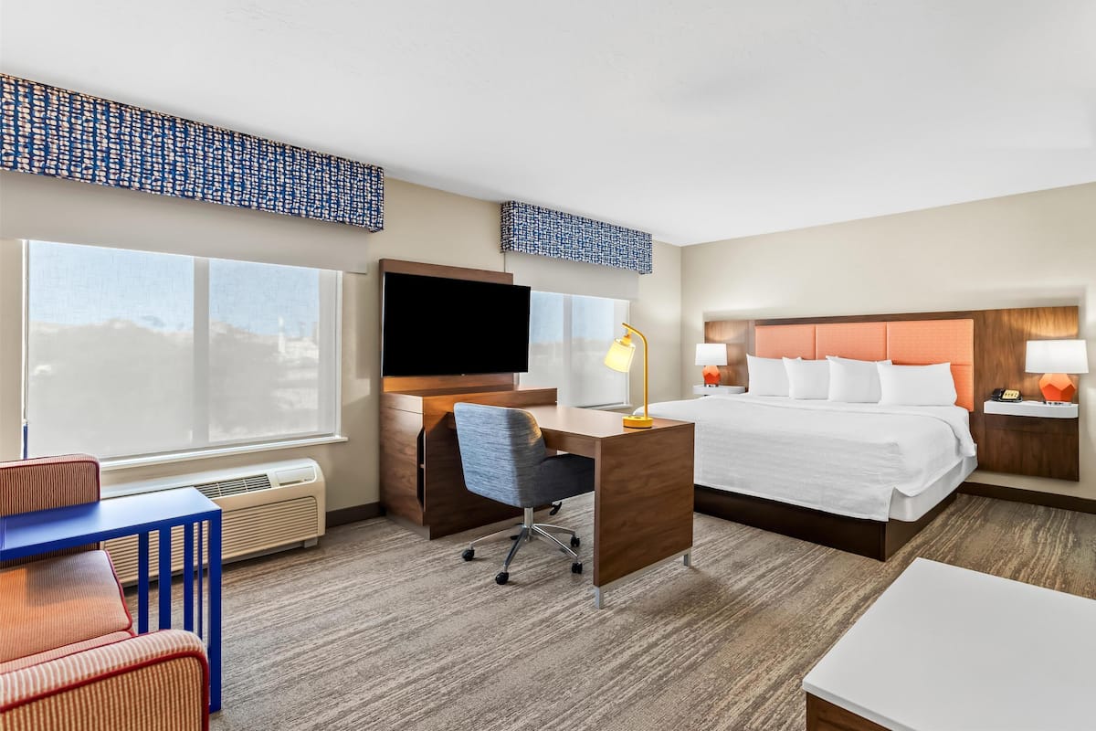 Habitación de hotel con cama, escritorio, silla, televisor y ventanas amplias.