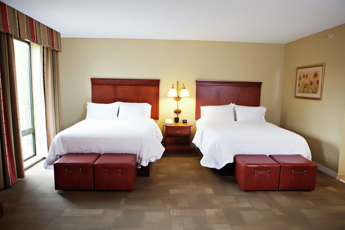 A hotel room with two beds, a nightstand, a lamp, and a window with curtains.