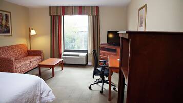 A hotel room with a bed, a sofa, a desk, a chair, an air conditioner, and a television.