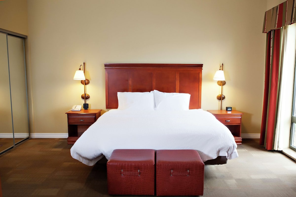 A hotel room with a large bed, two bedside tables with lamps, a wooden headboard, and a large window with curtains.